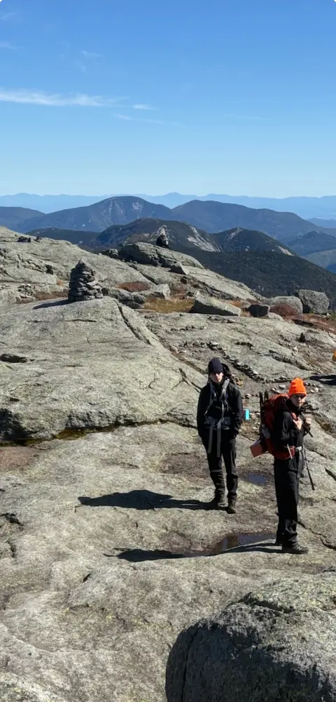 Two hikers standing on the bare granite summit, a cairn and rows of Adirondack peaks beyond.