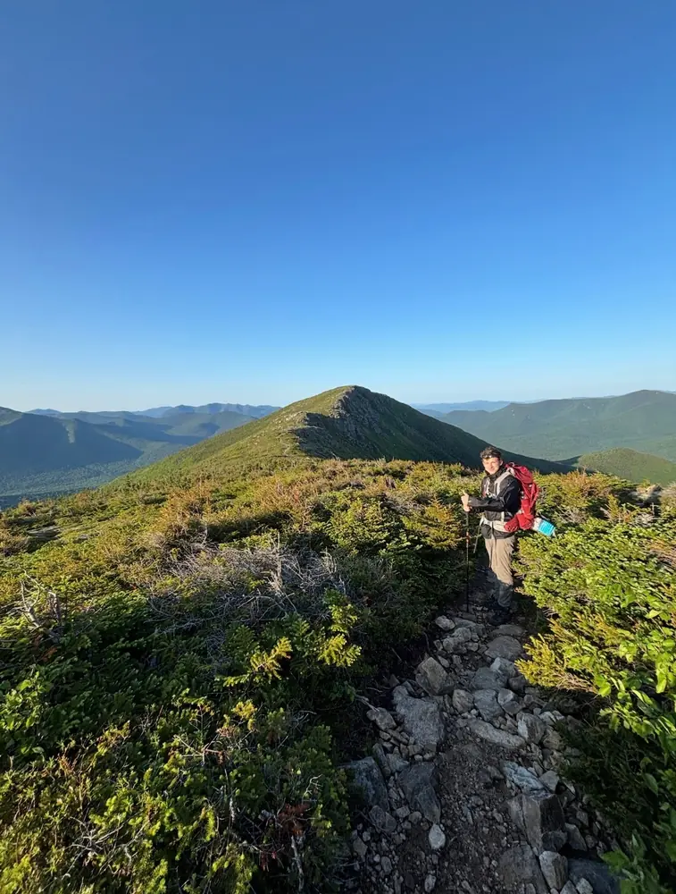 A hiker with a red pack and trekking poles on a rocky trail through low scrub, a bare ridge and blue mountains behind.